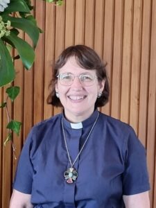 Woman, smiling. Wearing blue clerical shirt and cross.