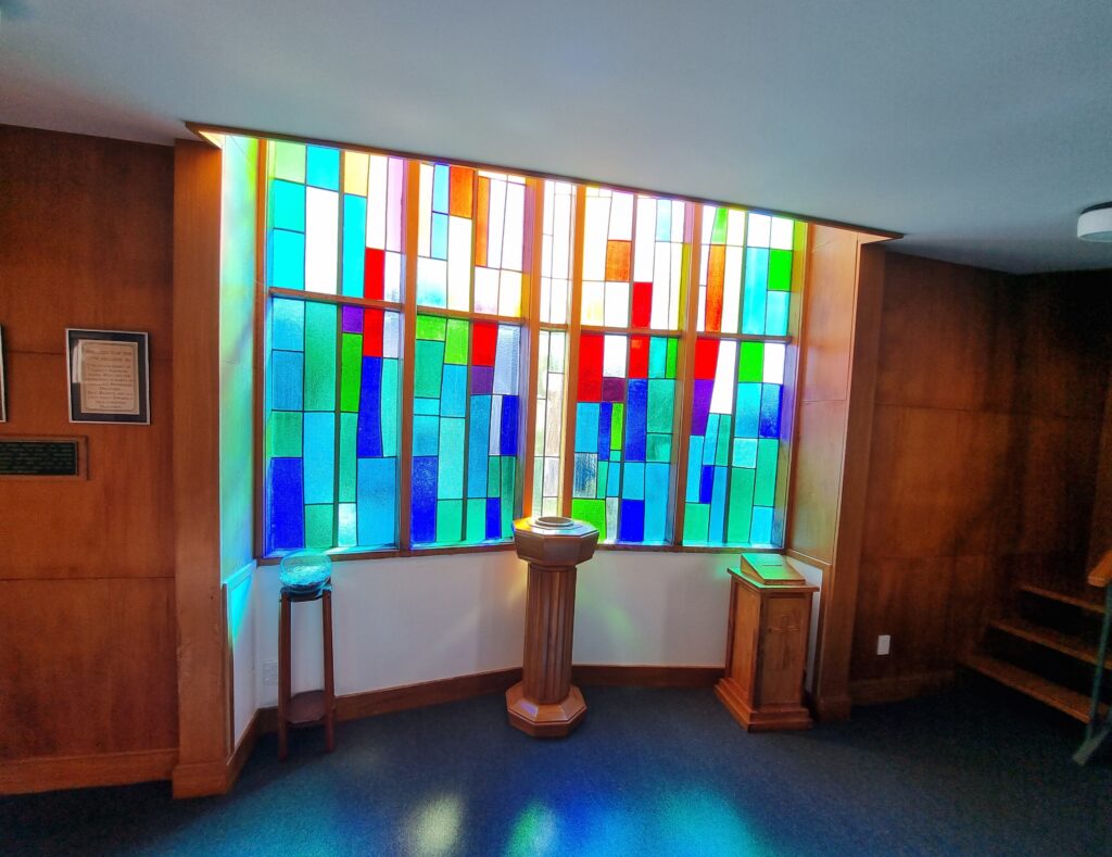 Multi-coloured window and the baptismal font in front; other furniture and internal stairs visible. Sunlight is streaming on to the carpet.