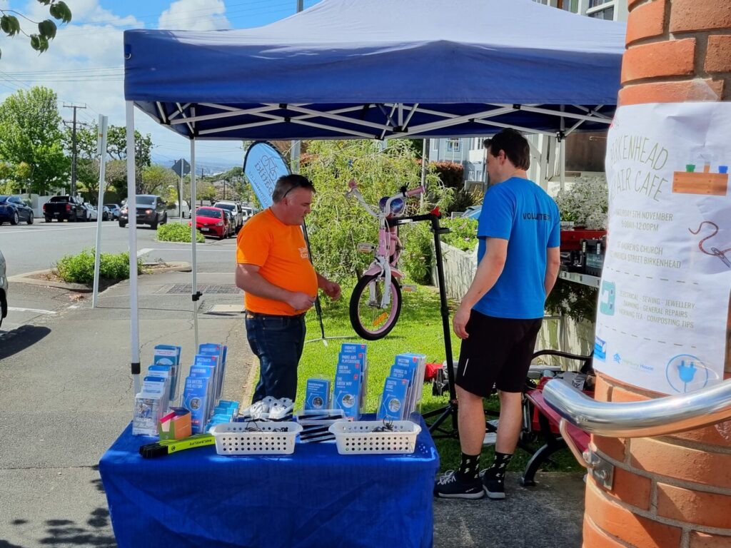 Two men working on a child's bike. A table of brochures in the foreground.