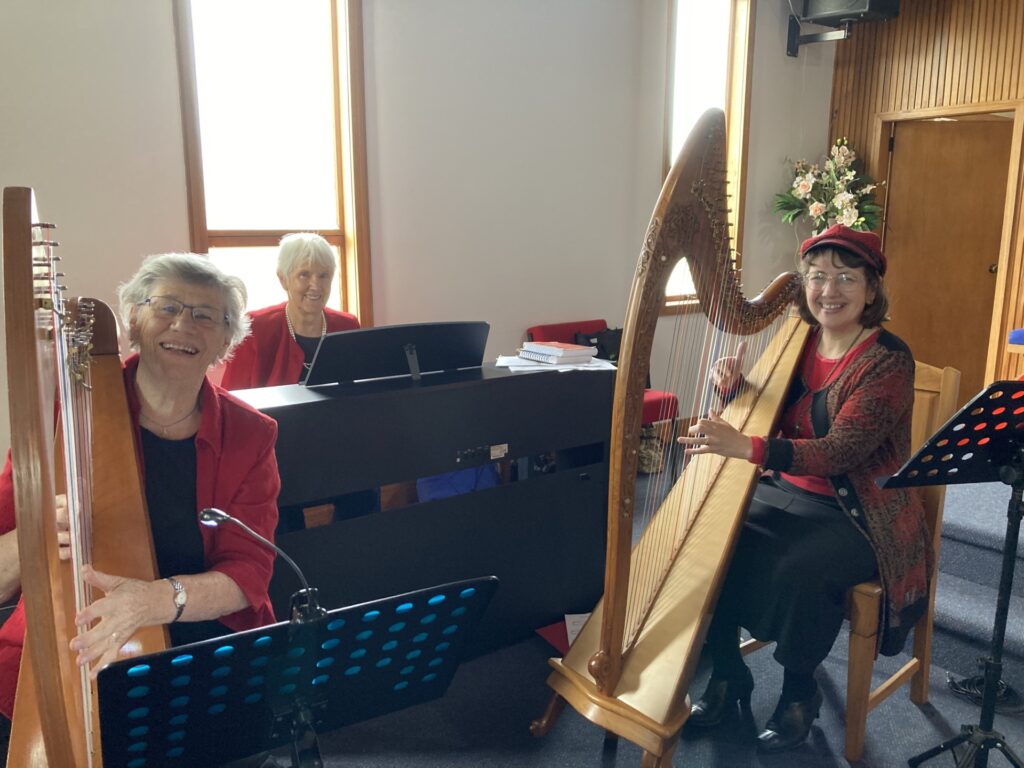 Keyboard player at the organ; two harp players with their harps. All three women are dressed in red and smiling. The harp players have their fingers on the harp strings.