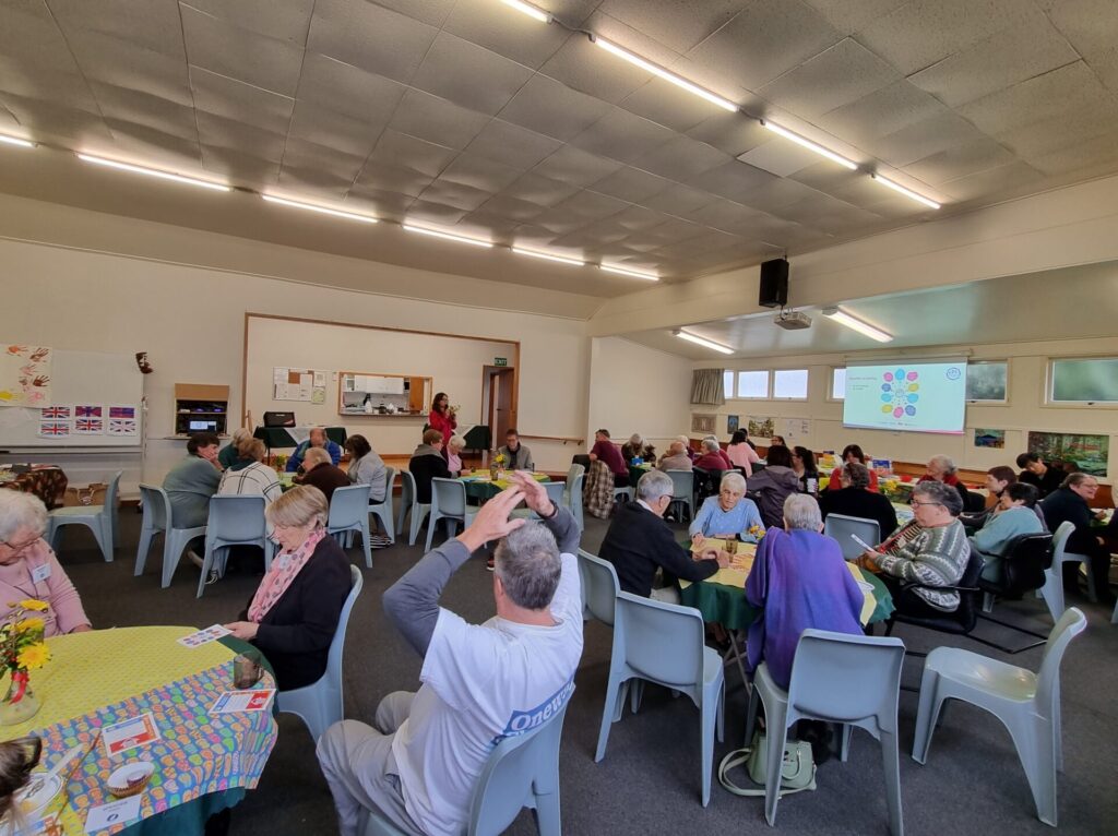 Hall of people sitting at round tables. They look at Neighbourhood Support material as a woman is speaking.