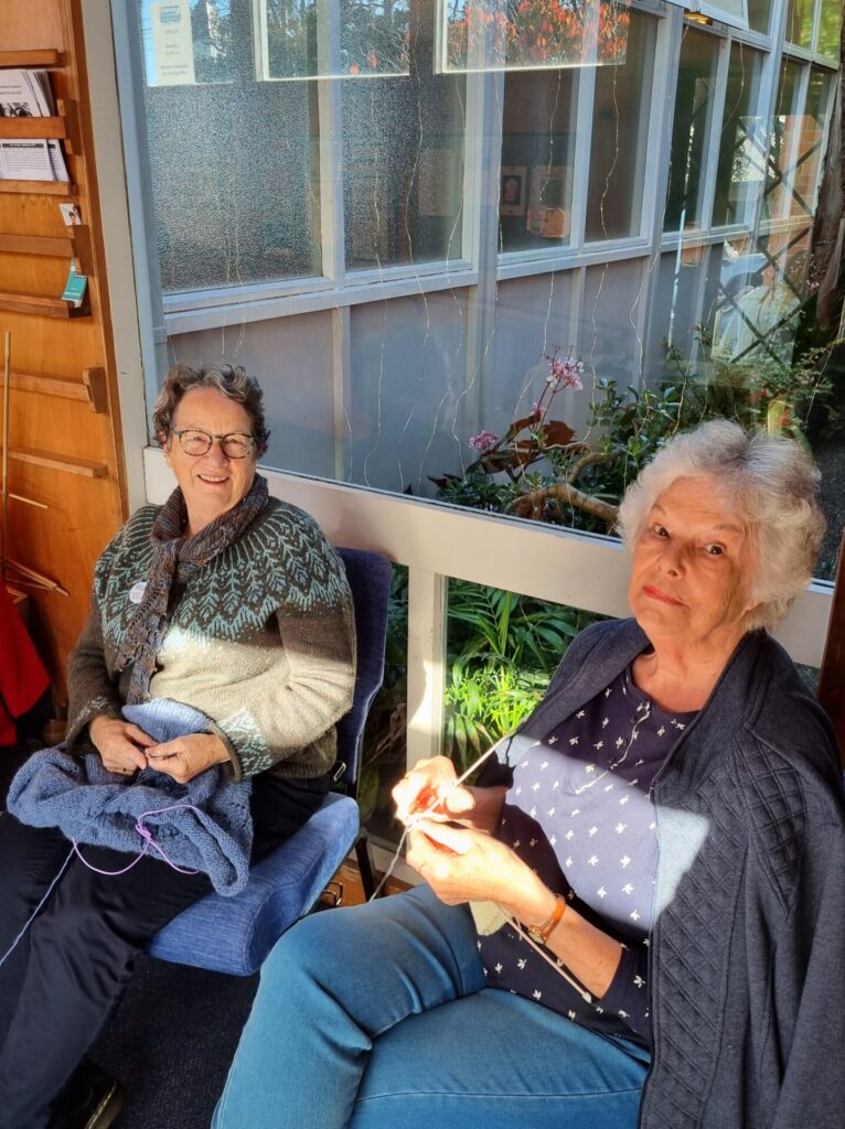 Two women knitting. The sun is shining on them. Garden visible in the background.