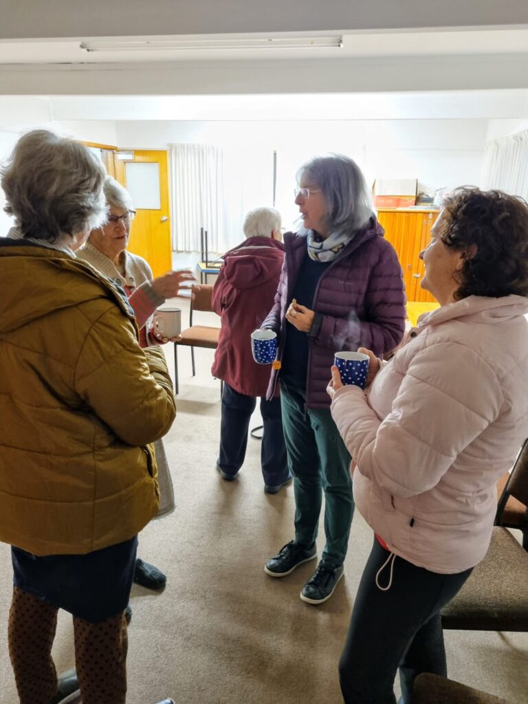 Group of four women are standing and talking; they are holding cups of tea.