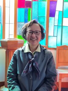 Woman sitting in the church. She is Korean; wears glasses and is smiling. Sun is streaming through the coloured window behind her.