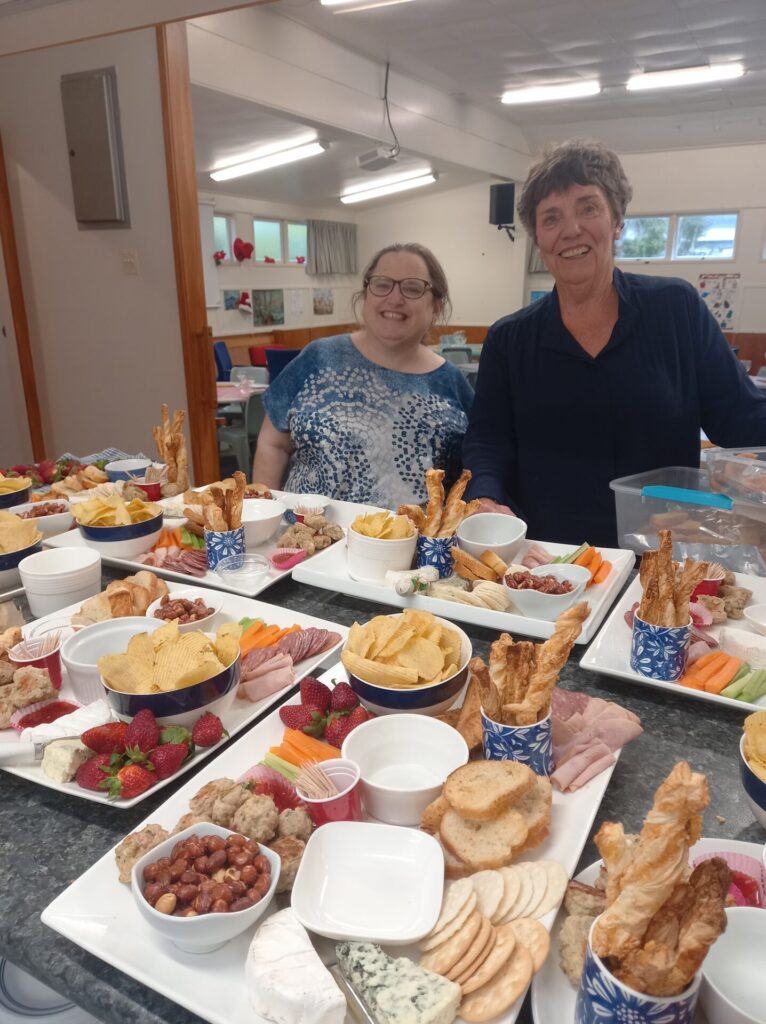 Two women are smiling at the camera as they eye up the platters they are arranging.