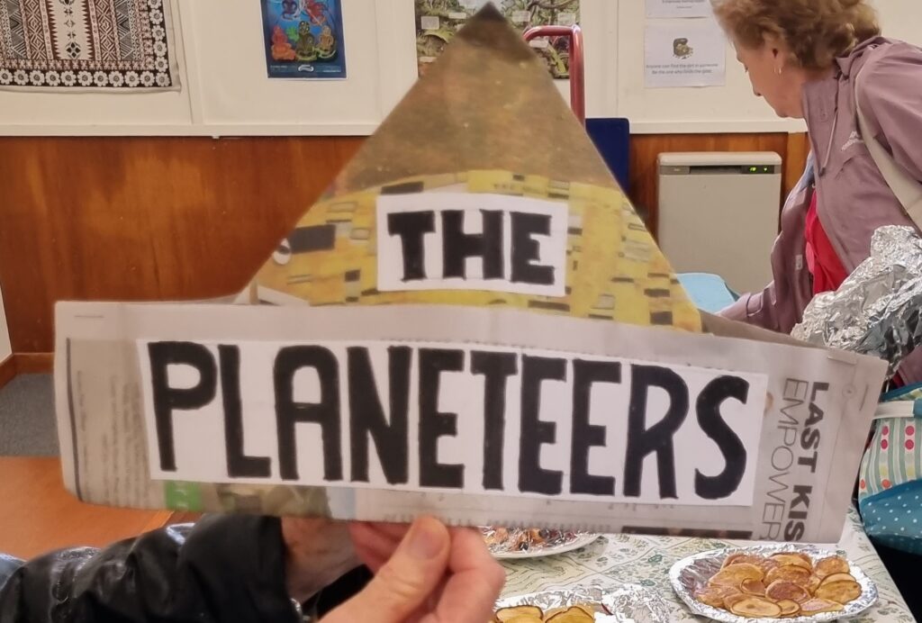 Folded newspaper hat with 'THE PLANETEERS' on front.