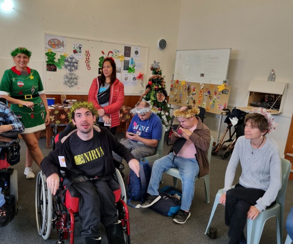 Assortment of people in Christmas clothing are seated in a decorated hall. Two people are in wheelchairs.