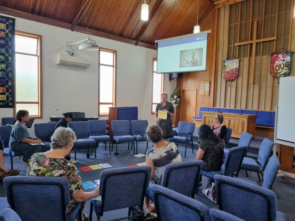 A woman presenting in the church, group of people listening. Coloured cards are laid out on the floor.