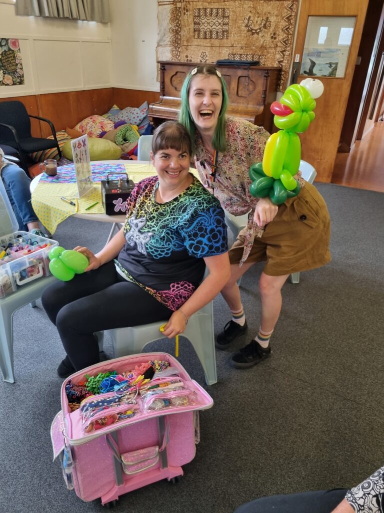 Two women smile enthusiastically at the camera. One is holding a large green frog made from balloons. Stacks of balloons ready for making into things. In the church hall - piano, cushions and tapa cloth visible in the background.