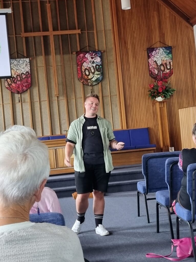 A man in his 30s speaks at the front of the church. Behind him are the banners that read 'peace,' 'love' and 'joy.'