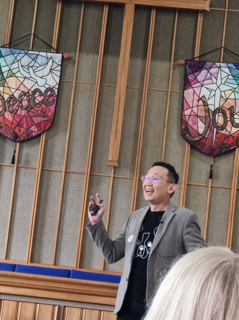 Asian man is looking upwards and laughing as he presents. He is speaking at the front of the church.