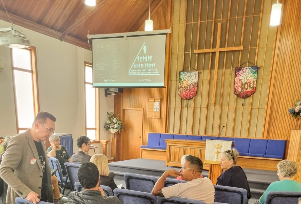 Diverse group of people seated and listening to one man speak. Ivan, the facilitator, is standing.