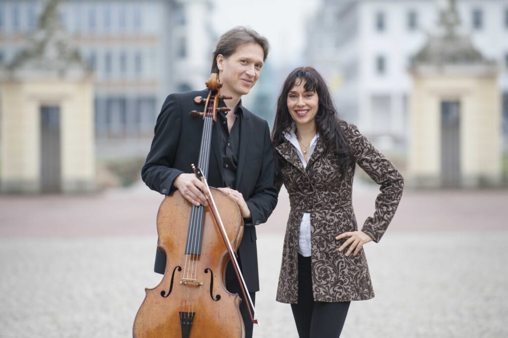 A man and a woman are outside, smiling at the camera. The man is holding a cello.