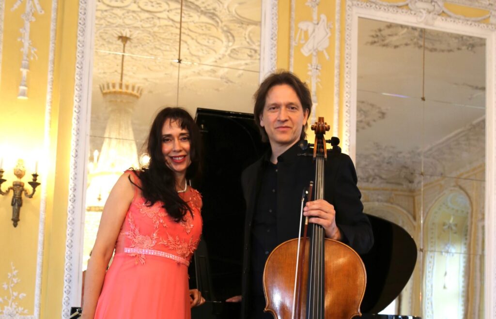 A man and a woman are in a grand room. They are smiling at the camera. The man is holding a cello. A grand piano is behind them.